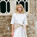 A woman with blonde hair stands in front of a stone wall and window, wearing a white short-sleeve button-up shirt dress with a beige sash tied at the waist.