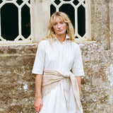 A woman with blonde hair stands in front of a stone wall and window, wearing a white short-sleeve button-up shirt dress with a beige sash tied at the waist.