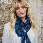 A woman with long blonde hair wearing a navy blue scarf with a bright blue paisley pattern and a decorative silver-toned brooch, posed against a textured stone wall.