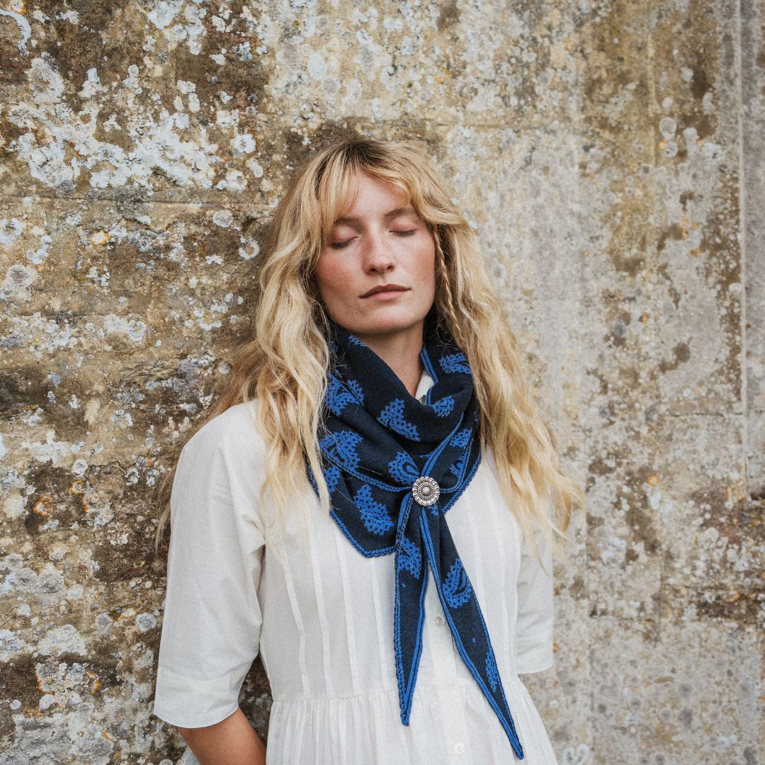 A woman with long blonde hair stands against a textured stone wall, wearing a white dress and a dark blue patterned scarf with a decorative silver-toned slider.
