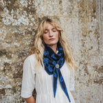 A woman with long blonde hair stands against a textured stone wall, wearing a white dress and a dark blue patterned scarf with a decorative silver-toned slider.