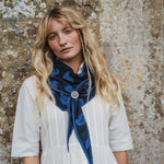 A woman with long blonde hair wearing a white dress and a navy blue patterned scarf with a silver-toned slider, standing against a textured stone wall.