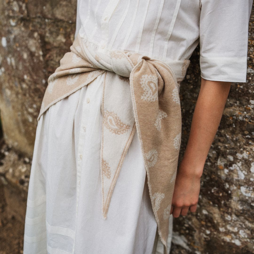 A person wearing a white dress tied at the waist with a beige paisley scarf, standing against a textured stone wall.