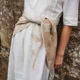 A person wearing a white dress tied at the waist with a beige paisley scarf, standing against a textured stone wall.