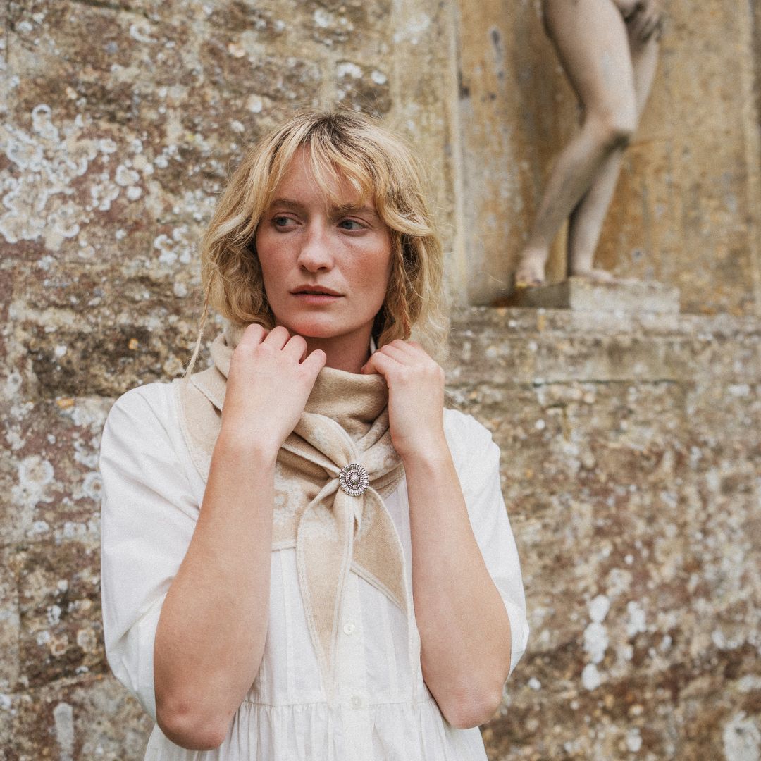 A woman with blonde hair stands in front of a textured stone wall, wearing a cream-colored dress and a beige scarf tied at the neck with a decorative brooch.