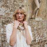 A woman with blonde hair stands in front of a textured stone wall, wearing a cream-colored dress and a beige scarf tied at the neck with a decorative brooch.