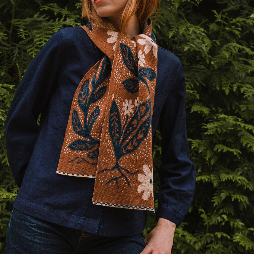 person in the woods wearing a brown knitted botanical scarf with cream and blue floral pattern by cotswold knit.
