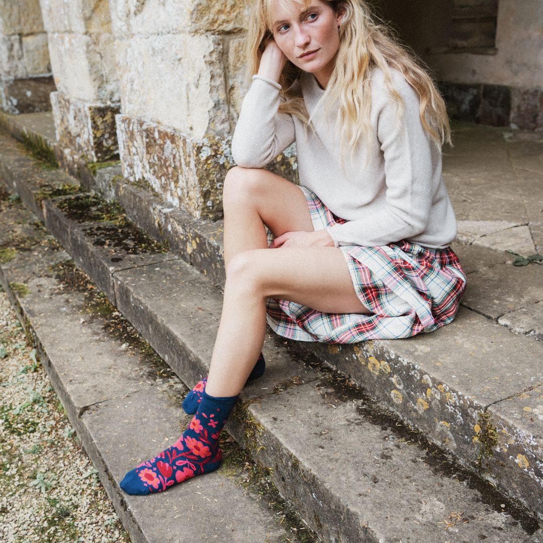 A woman sits on stone steps wearing a cream sweater, a red and white plaid skirt, and navy socks with a red and pink abstract pattern.