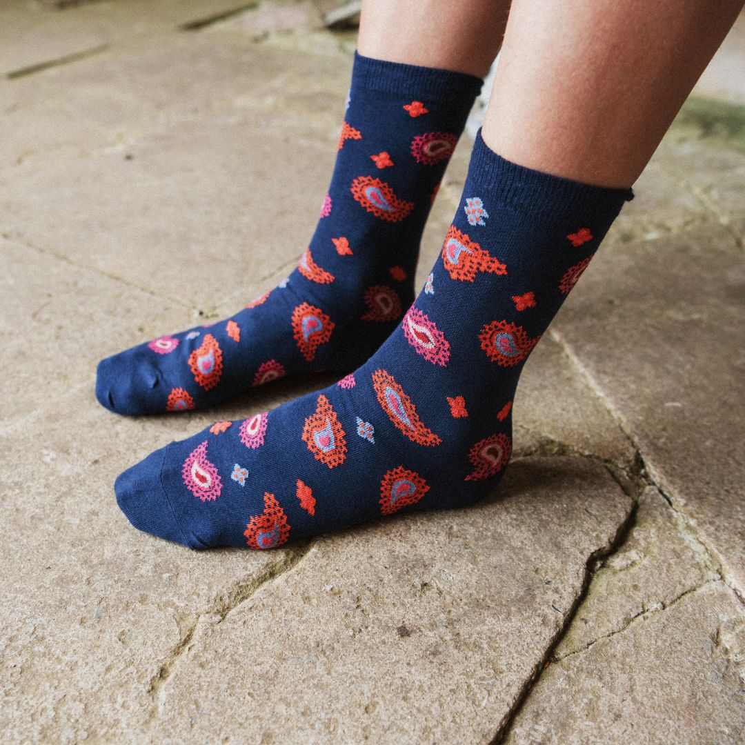 A close-up of a person's feet wearing navy blue crew socks with a red, orange, and pink paisley pattern, standing on a stone pavement surface.