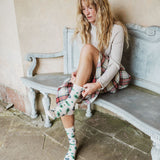 A woman sits on a weathered bench wearing a cream sweater, a red and green plaid skirt, and white crew socks with a green pine tree pattern.