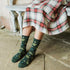 A person seated on a stone bench wearing dark green crew socks with a light green paisley and clover pattern, paired with a red, green, and white plaid skirt.
