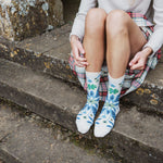 A person sitting on stone steps wearing white crew socks with blue and green floral patterns, paired with a red and green plaid skirt and a light gray sweater.