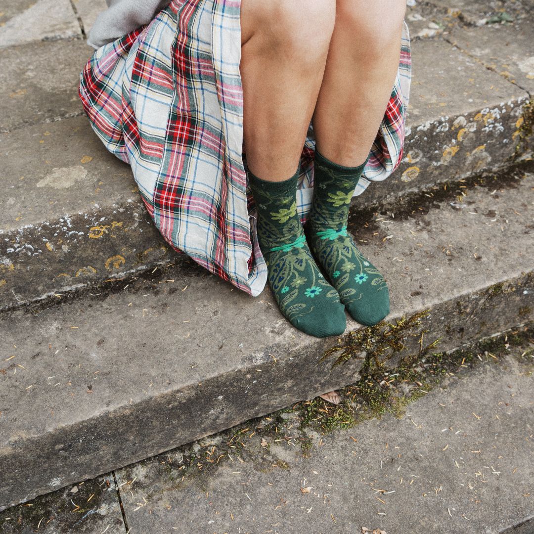 A person sitting on concrete steps wearing dark green floral socks and a red, white, and green plaid skirt.