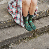 A person sitting on concrete steps wearing dark green floral socks and a red, white, and green plaid skirt.