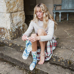 A woman sits on stone steps wearing a cream knit sweater, a red and green plaid skirt, and white socks with a blue tropical leaf pattern. She is holding one of the socks up to show the design.