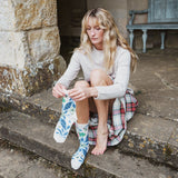 A woman sits on stone steps wearing a cream knit sweater, a red and green plaid skirt, and white socks with a blue tropical leaf pattern. She is holding one of the socks up to show the design.