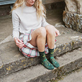 A woman sits on stone steps wearing a light beige sweater, a red and green plaid skirt, and dark green patterned socks.