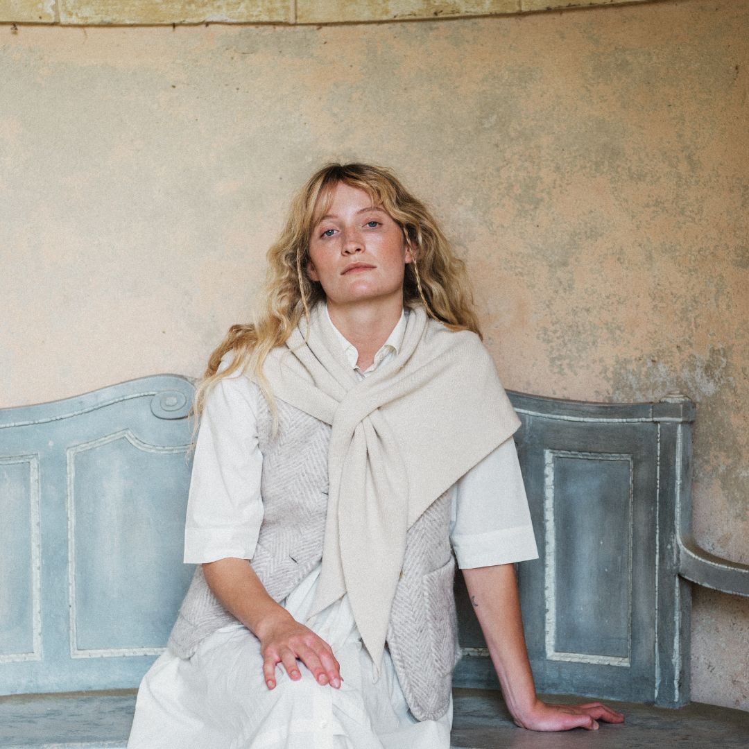 A woman with blonde hair sits on a distressed blue bench against a textured beige plaster wall, wearing a white shirt, a light beige knit vest, and a cream-colored scarf.