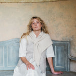 A woman with blonde hair sits on a distressed blue bench against a textured beige plaster wall, wearing a white shirt, a light beige knit vest, and a cream-colored scarf.