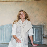 A woman with blonde hair sits on a distressed blue bench against a textured beige plaster wall, wearing a white shirt, a light beige knit vest, and a cream-colored scarf.