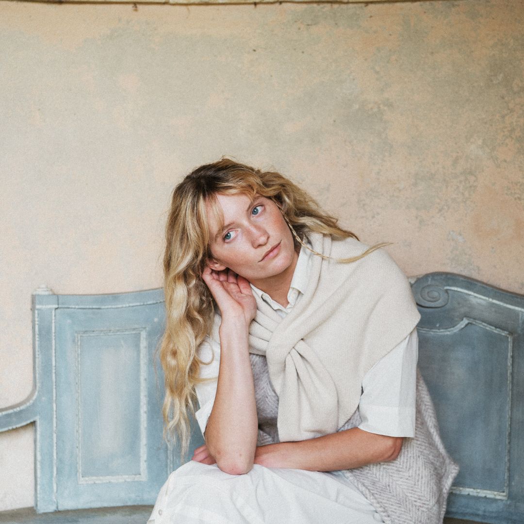 A woman with blonde hair sits on a distressed blue wooden bench against a textured beige wall, wearing a cream knit shawl over a white collared shirt and light-colored dress.