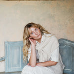 A woman with blonde hair sits on a distressed blue wooden bench against a textured beige wall, wearing a cream knit shawl over a white collared shirt and light-colored dress.