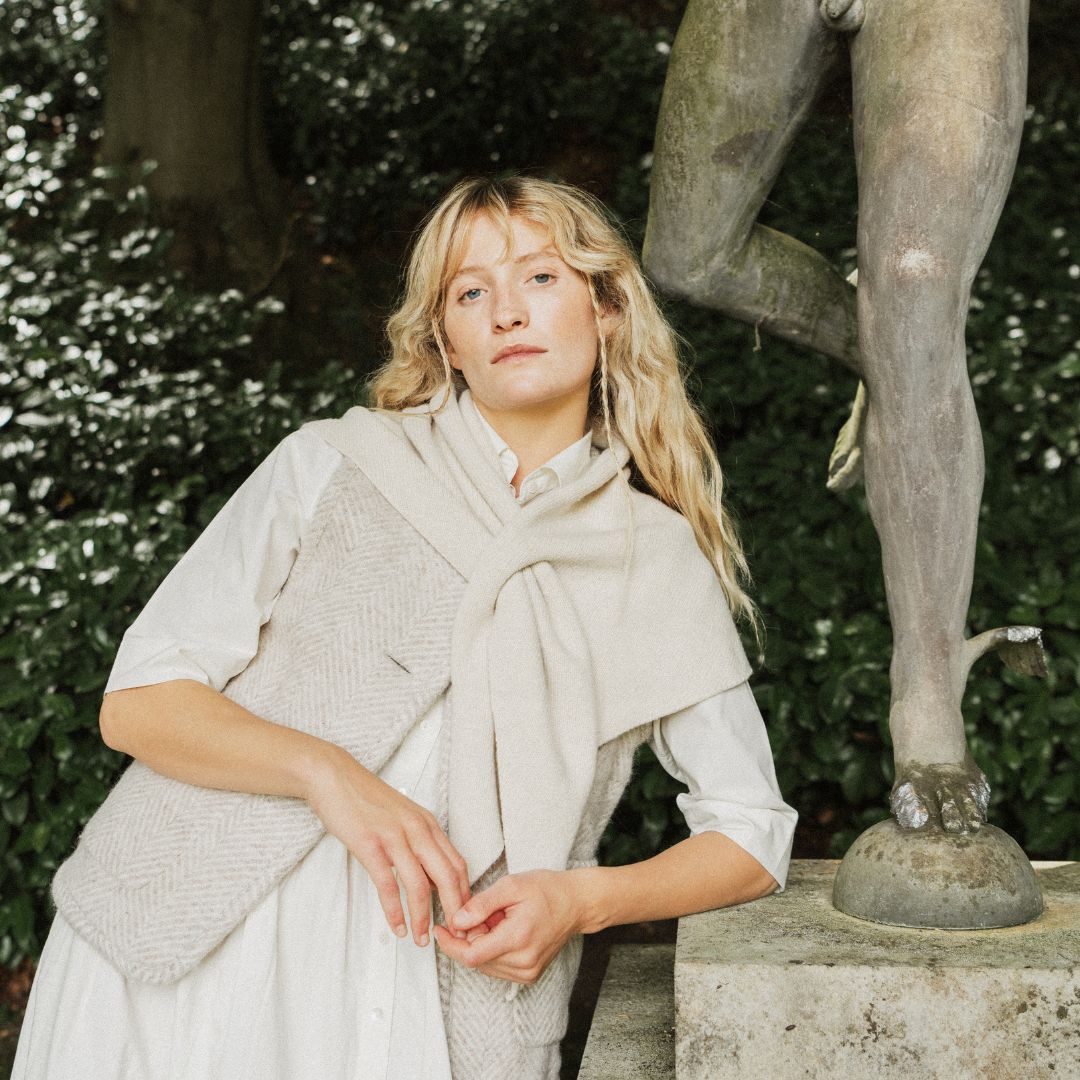 A woman wearing a cream-colored knit vest and matching scarf stands outdoors next to a stone statue, with green foliage in the background.