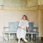 A woman wearing a long white dress, white socks, and a light beige shawl sits on a weathered blue-gray bench against a textured beige stone wall.
