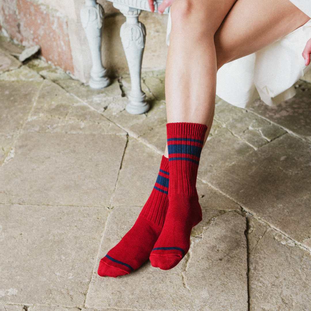 A person sitting on a stone floor wearing red crew socks with navy blue stripes near the top and toes.