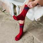 A person sitting on a stone floor wearing red crew socks with navy blue stripes near the top. One hand is adjusting the sock on the leg.