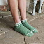 A person wearing ribbed ankle socks in sage green with white stripes, standing on a stone floor.