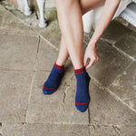 A person sitting on a stone bench wearing navy blue ribbed ankle socks with red stripes at the cuff and toe, adjusting one sock with their hand.