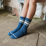 A person wearing blue crew socks with two white horizontal stripes near the top, shown from the knees down while sitting on a stone floor.