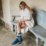 A woman sits on a weathered blue bench in a rustic indoor setting, wearing a loose white shirt dress and blue crew socks with white stripes.