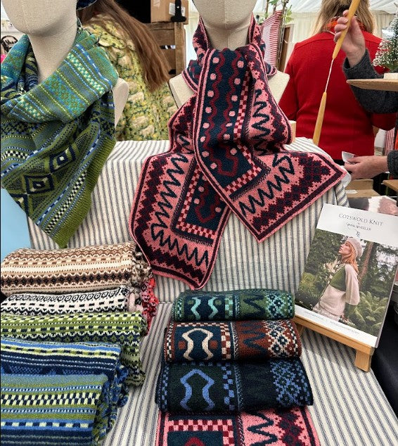 A display of multiple knitted scarves with intricate geometric patterns, shown on mannequins and folded on a striped table. The scarves feature colorways including green, pink, navy, and multicolor patterns.