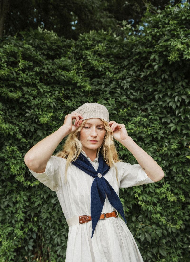 A woman stands outdoors in front of a leafy green wall, wearing a light-colored dress, a navy blue neck scarf, a beige beret, and a brown belt.