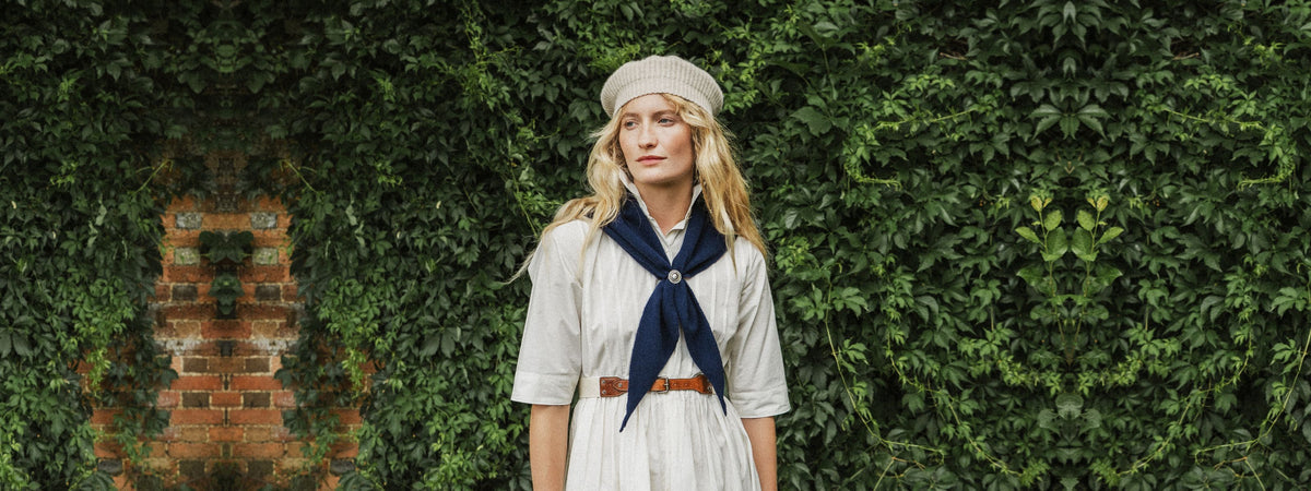 A woman stands outdoors in front of a leafy green wall, wearing a light-colored dress, a navy blue neck scarf, a beige beret, and a brown belt.