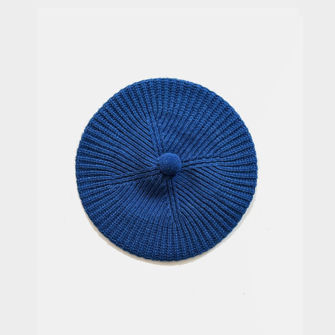A blue knit beret with a textured radial pattern and a small matching knit pom-pom on top, photographed flat against a light gray background.