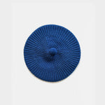 A blue knit beret with a textured radial pattern and a small matching knit pom-pom on top, photographed flat against a light gray background.