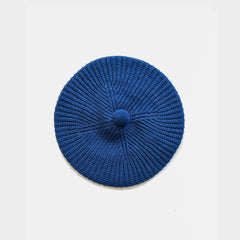 A blue knit beret with a textured radial pattern and a small matching knit pom-pom on top, photographed flat against a light gray background.