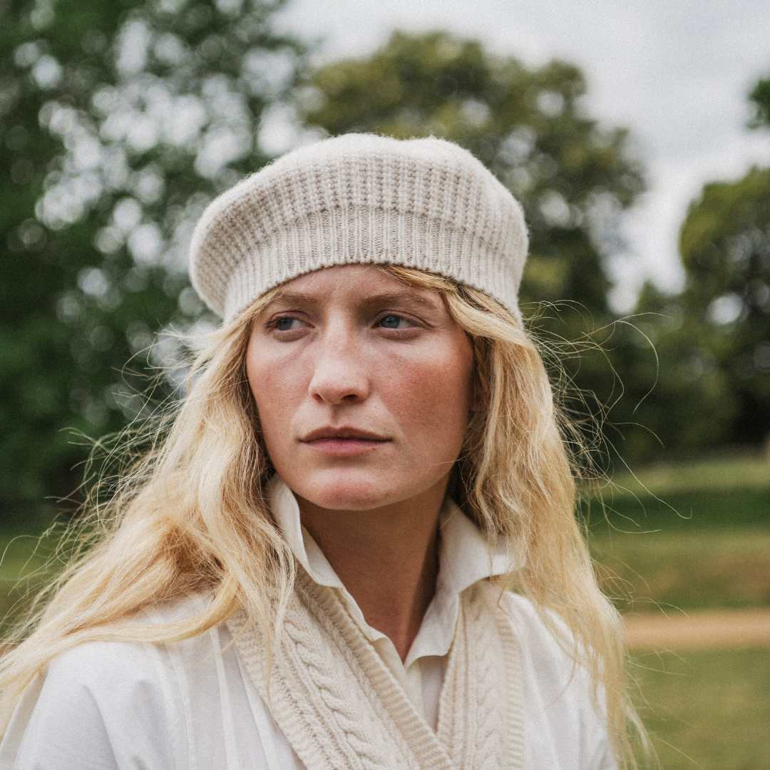 A woman outdoors wearing a cream-colored ribbed knit beret and a matching cream cable-knit sweater over a white collared shirt, with a blurred green forest background.