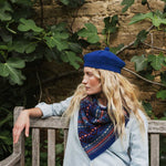 A woman with long blonde hair sits on a wooden bench outdoors, wearing a bright blue knit beanie, a blue and red patterned scarf, and a light blue jacket. She is posed against a rustic stone wall with green fig leaves.