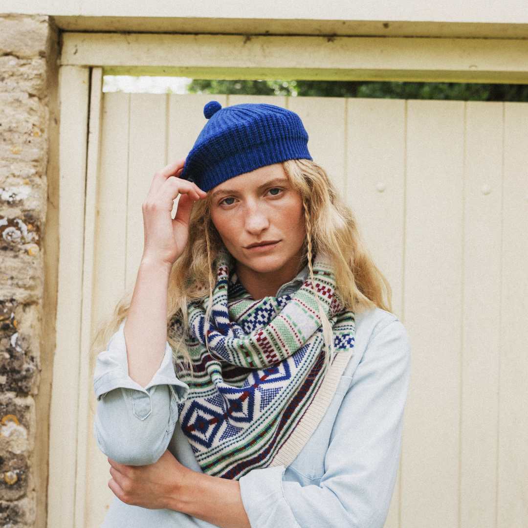 A woman wearing a blue knit beret with a pom-pom and a multicolor geometric patterned scarf, standing in front of a cream-colored wooden door.