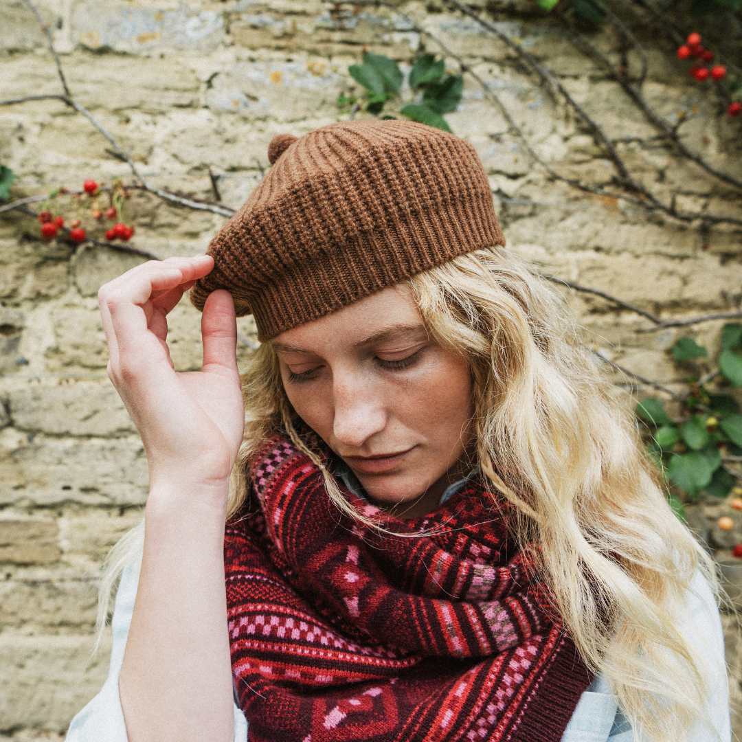 A woman wearing a brown ribbed knit beret and a red patterned knit scarf, posing outdoors against a rustic stone wall with climbing plants and red berries.