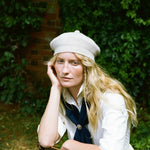 A woman with long blonde hair wearing a white knit beret, white shirt, and dark navy necktie, posing outdoors against a background of green foliage and a brick wall.
