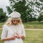 A woman outdoors wearing a cream-colored ribbed knit beret and a matching cream knit cardigan over a white dress, standing in a grassy park with trees in the background.