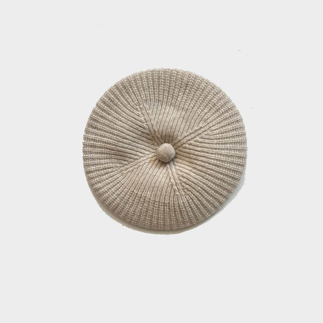 A round, ribbed knit cap in a light beige or oatmeal color, featuring a central button detail, photographed from directly above on a light gray background.