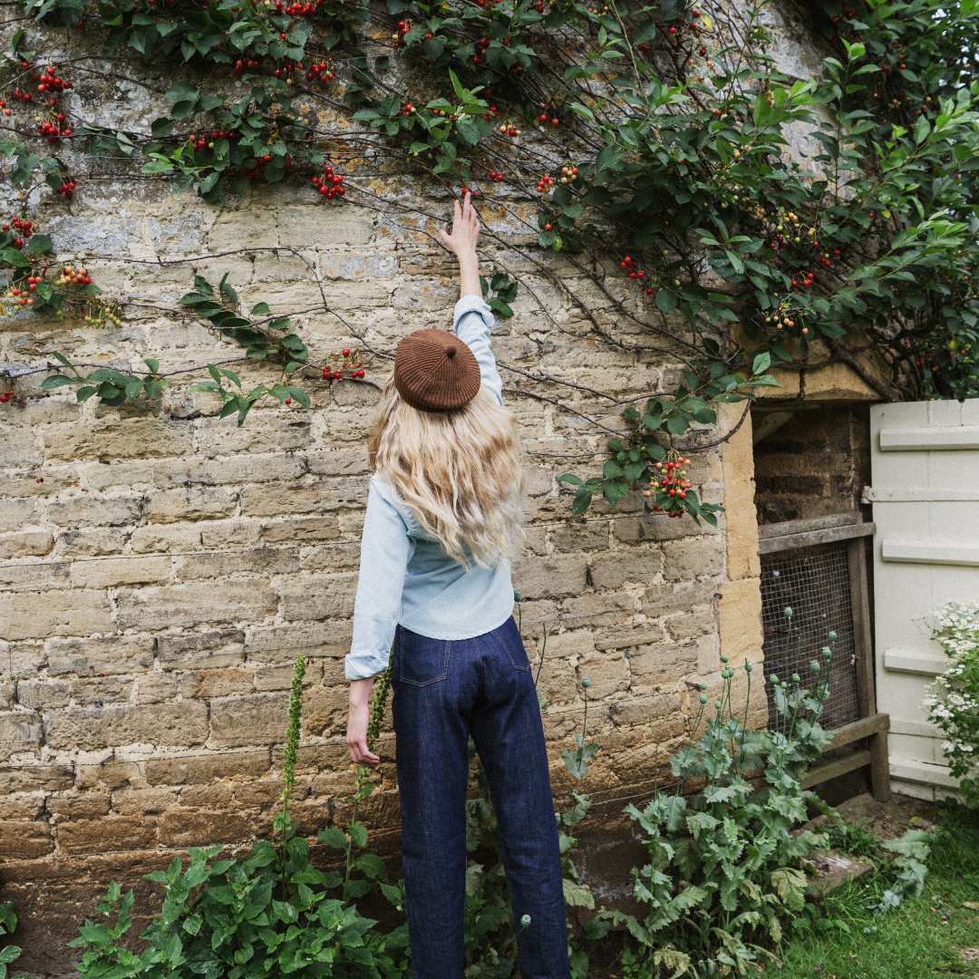 A woman seen from behind wearing a brown beret, light blue denim jacket, and dark blue jeans, standing in front of a rustic stone wall covered in green ivy and small red berries.