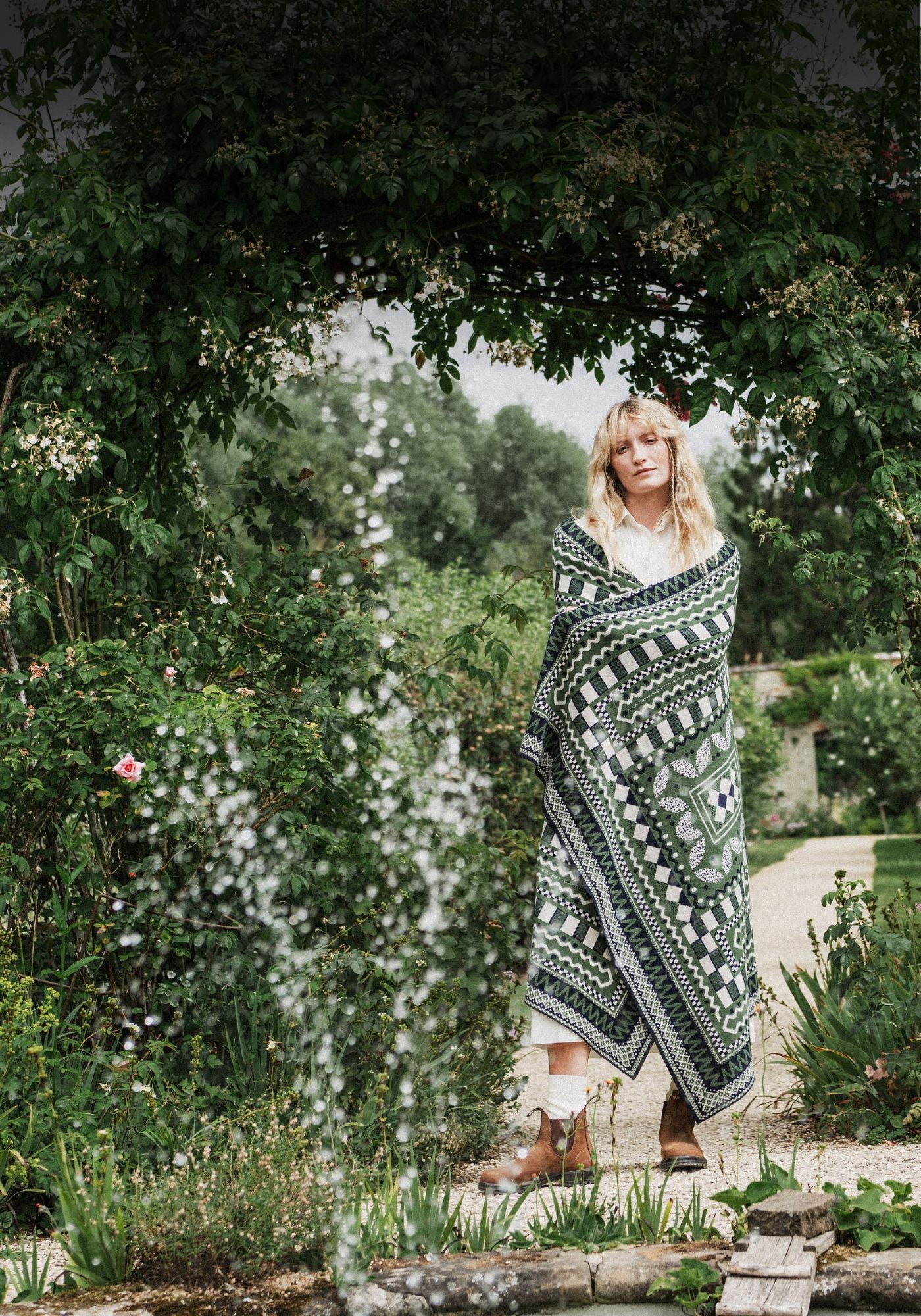 A woman standing in a garden pathway wrapped in a large green and white geometric patterned blanket, with lush foliage and a fountain in the foreground.