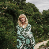 A woman with blonde hair stands outdoors in a garden setting, wrapped in a dark green knit blanket featuring a large-scale mint green and white floral pattern.